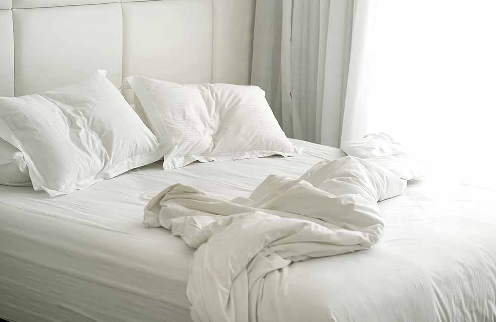 Ein benutztes Doppelbett mit weißer Bettwäsche steht mit zurückgeschlagenen Decken vor einem bodentiefen Fenster.