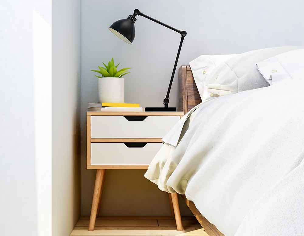 Ein kleiner hölzerner Nachhtisch mit 2 weißen Schubladen steht neben einem Bett. Auf ihm befindet sich eine Nachhtischlampe, eine Grünpflanze und 2 Bücher.