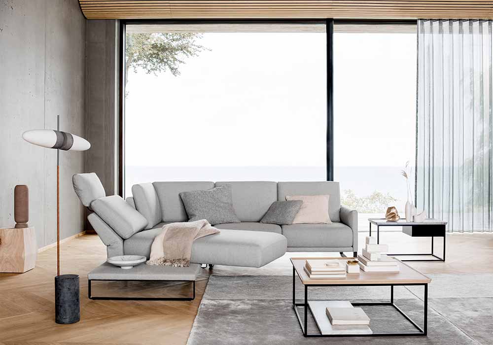 Modernes graues Ecksofa steht in einem Wohnzimmer vor großen bodentiefen Fenstern.
