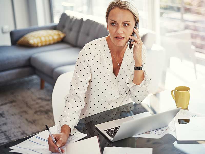 Eine Frau sitzt in Ihrem Wohnzimmer am Tisch vor ihrem Laptop und telefoniert