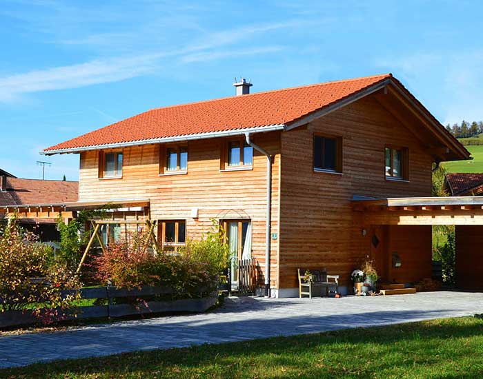 Ein Holzhaus mit Holzpergola über der Terrasse und holzüberdachten Stellplätzen steht auf einem sonnigen Grundstück